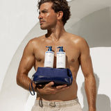 A shirtless man holds a navy blue leather toiletry bag with two white pump bottles from the brand RAN, set against a clean white architectural background.